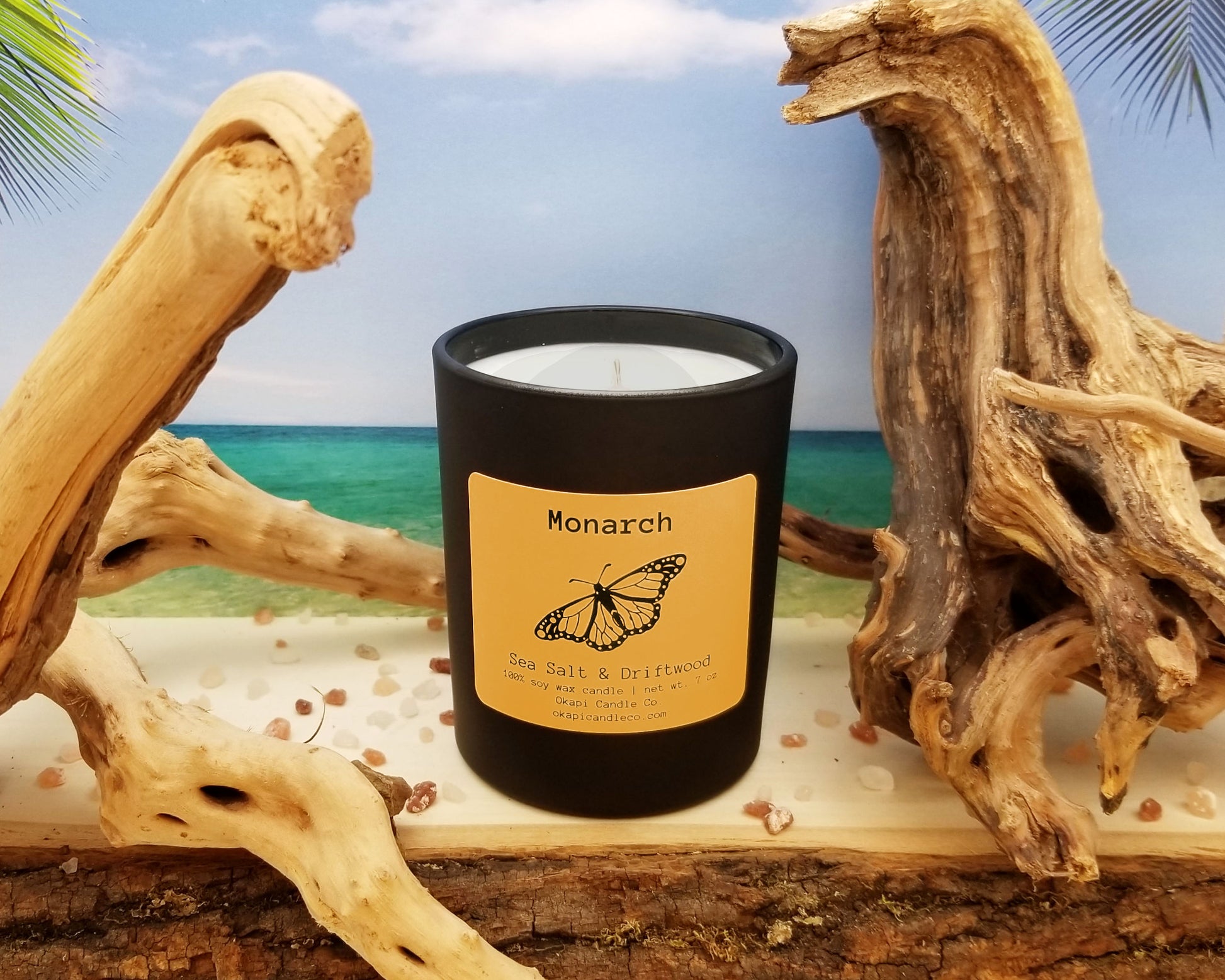 Gnarled driftwood and pink sea salt surround a candle in a tasteful black tumbler with an orange label that features a small but detailed monarch design.