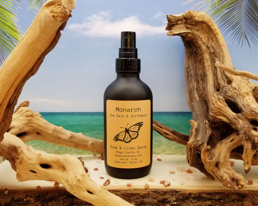 Gnarled driftwood and pink sea salt surround a room spray in a tasteful black glass spray bottle with an orange label that features a small but detailed monarch design.