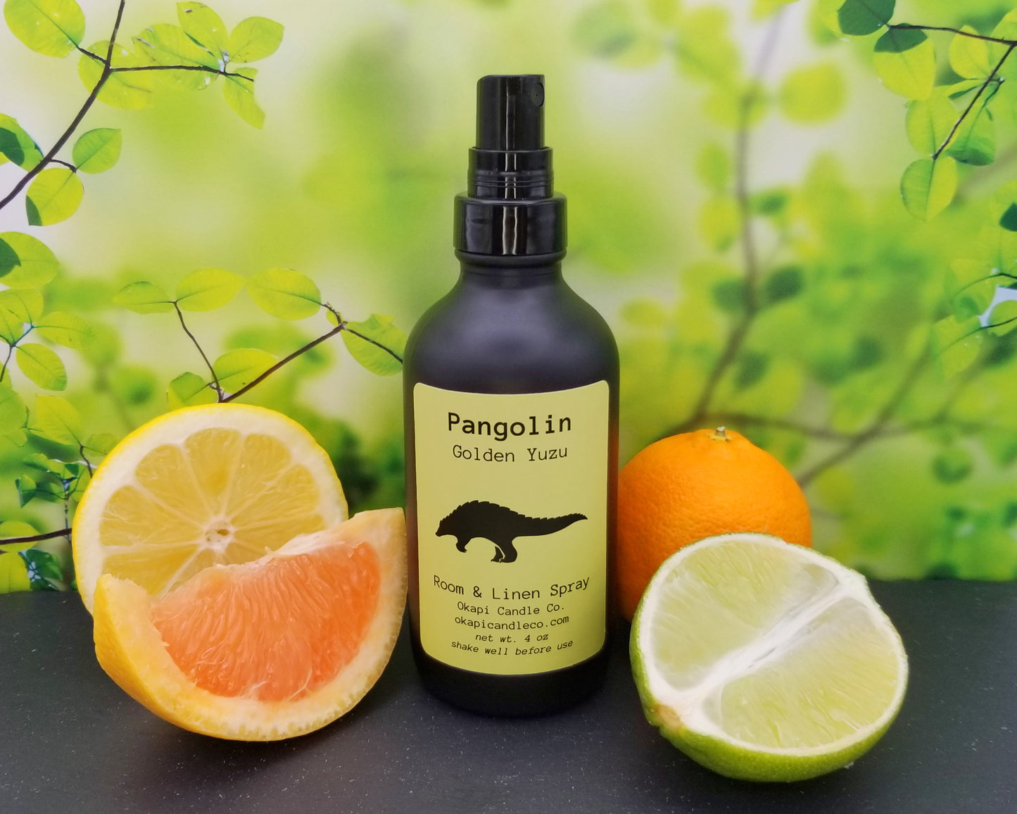 A room spray in a black glass spray bottle, surrounded by various citrus fruits, sits in front of a bright, leafy backdrop. The candle bears a yellow label with an illustration of a pangolin. 